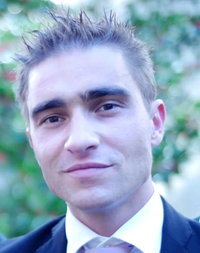 Ein Mann mit kurzem Haarschnitt und einem Lächeln im Gesicht, trägt einen schwarzen Anzug und ein weißes Hemd. Er scheint draußen zu stehen, mit grünem Laub im Hintergrund.