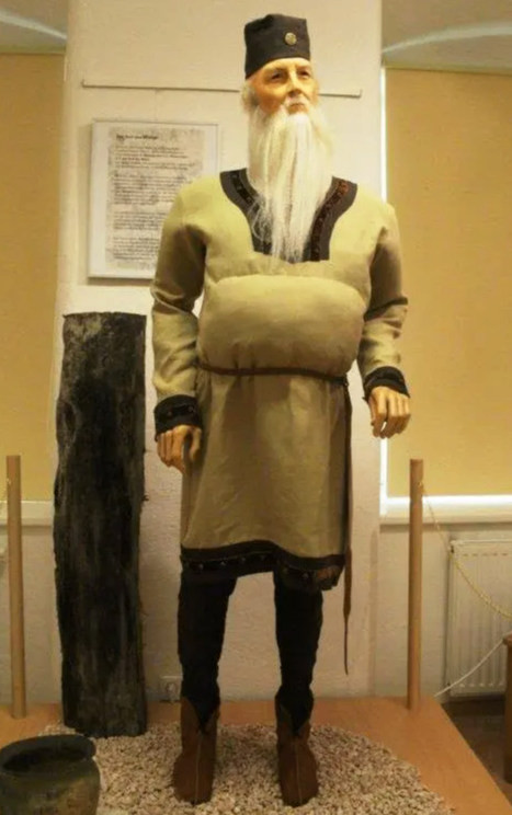 Eine Wachsfigur in traditioneller Kleidung steht in einem Museum neben einem großen Baumstamm und einem Blatt Papier an der Wand.