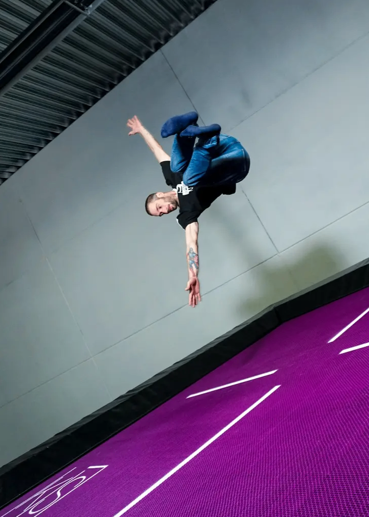 Ein Mann in einem schwarzen T-Shirt und blauen Jeans führt einen akrobatischen Sprung in der Luft auf einem Trampolin aus.