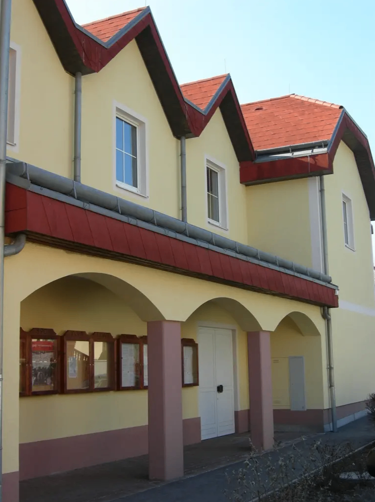 Ein Gebäude mit rotem Dach und gelben Wänden. Es hat drei gewölbte Eingänge, drei Fenster und drei kleine Holztafeln mit angehängten Papieren.
