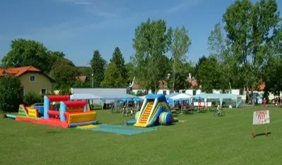 Ein Spielplatz mit aufblasbaren Rutschen und Hüpfburgen auf einer grünen Wiese mit Bäumen und Häusern im Hintergrund.