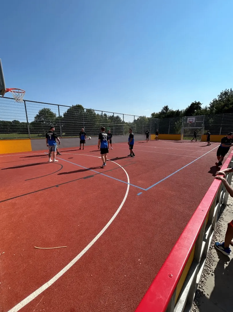 Eine Gruppe von Menschen spielt Basketball auf einem Platz mit roten und gelben Markierungen. Ein Zaun umgibt den Platz.
