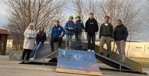 Eine Gruppe von Menschen posiert für ein Foto in einem Skatepark, auf Graffiti-bemalten Rampen stehend. Im Hintergrund sind kahle Bäume und ein Gebäude zu sehen.