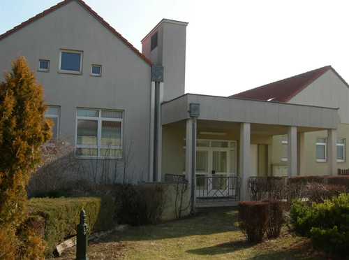 Ein modernes Haus mit grauer Außenfassade und rotem Dach, ausgestattet mit mehreren Fenstern, einer Glastür und einer kleinen Veranda mit Metallgeländer.