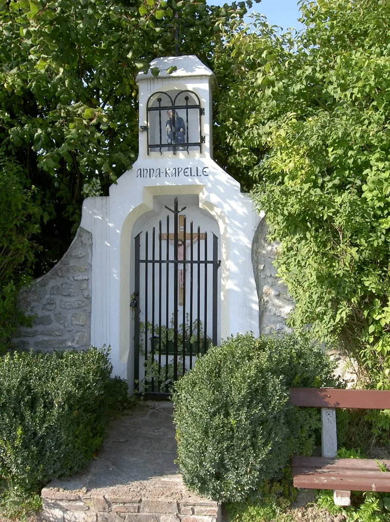 Eine weiße Kapelle mit einer Statue einer Person im Fenster und einem Kreuz über der Tür. Die Kapelle ist von grünen Pflanzen und Büschen umgeben.