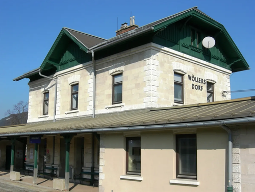 Das Wollers Dorf Bahnhofsgebäude hat eine beige Fassade mit grünen Verzierungen, mehrere Fenster und eine Satellitenschüssel auf dem Dach. Bänke stehen unter dem Vordach auf der linken Seite.