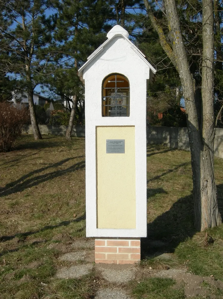 Eine kleine weiße und gelbe Struktur mit einem Glasfenster und einem Kreuz oben steht in einem Grasbereich mit Bäumen und einer Mauer.