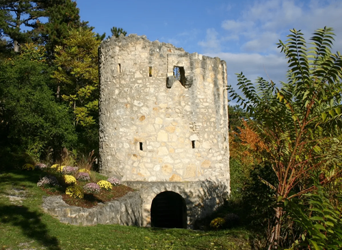Ein teilweise zerfallener Steinturm steht in einem Garten. Der Turm hat einen kleinen Eingang auf dem Boden, mit mehreren Löchern und einem zerbrochenen Fenster darüber. Grüner Rasen und bunte Blumen umgeben den Turm, und Bäume mit grünen und gelben Blättern sind im Hintergrund.