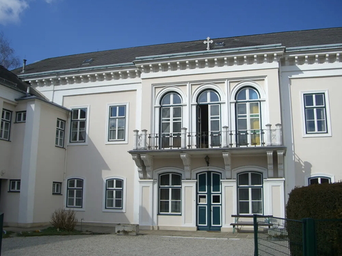 Ein großes weißes Gebäude mit Rundbogenfenstern und einem Balkon im zweiten Stock. Es hat eine grüne Tür und eine Bank davor.