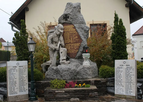 Ein Denkmal zeigt eine Skulptur eines Soldaten mit Gewehr und Helm, umgeben von Tafeln mit Namen und Daten. Die Umgebung umfasst einen Steinsockel mit einer Laterne, Grünpflanzen und ein Gebäude im Hintergrund.