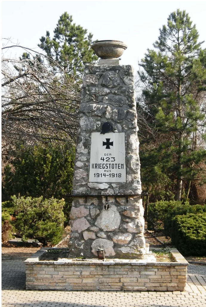 Ein Steinmonument mit einem Kreuz und einer Tafel, die 423 Kriegstoten 1914-1918 liest, steht in einem Park. Das Denkmal ist von Büschen und Bäumen umgeben.