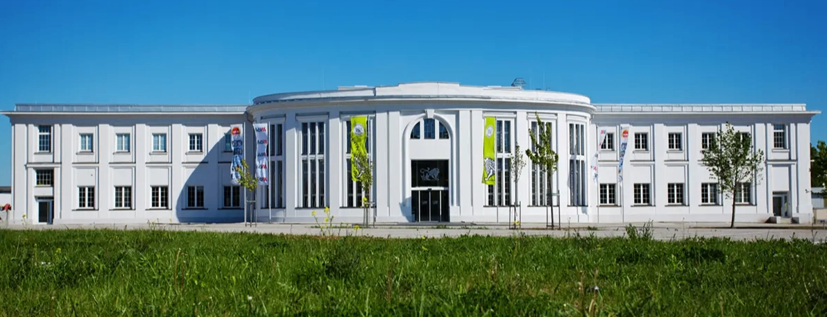 Ein modernes weißes Gebäude mit Rundbogenfenstern und grünen Bannern steht vor einer Rasenfläche unter einem blauen Himmel.
