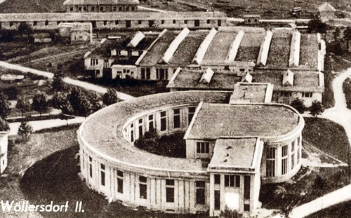 Eine Schwarz-Weiß-Luftaufnahme eines kreisförmigen Gebäudes mit vielen Fenstern und mehreren Gebäuden im Hintergrund.