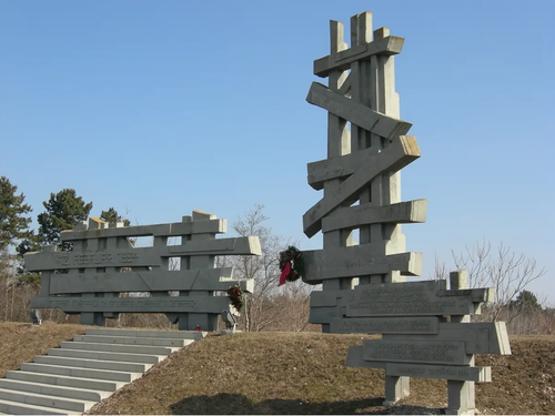 Ein großes Betondenkmal mit mehreren Tafeln steht unter klarem Himmel auf einem Hügel. Das Denkmal hat ein Zickzack-Muster und ist mit zwei Kränzen geschmückt.