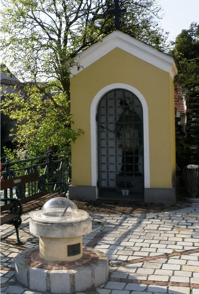 Eine kleine gelbe Kapelle mit einem Metallfenster und einer Tür steht auf einem Ziegelweg. Hinter der Kapelle ist ein Baum und ein Zaun zu sehen.
