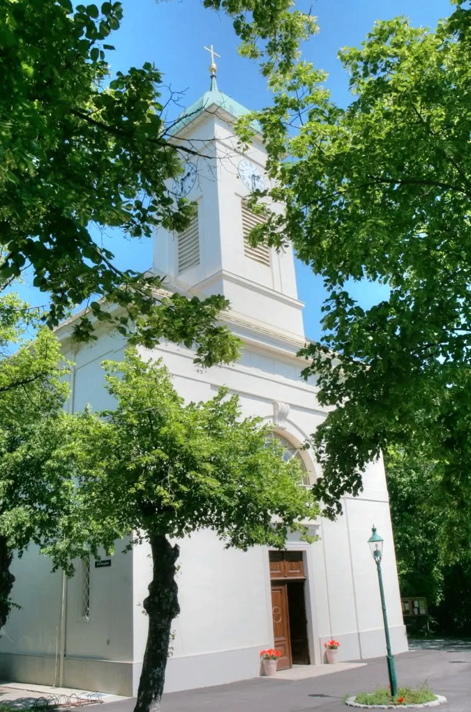 Eine weiße Kirche mit einem Glockenturm und einer grünen Straßenlampe ist von üppigen Bäumen umgeben. Die Kirche hat eine Holztür und Bogenfenster.