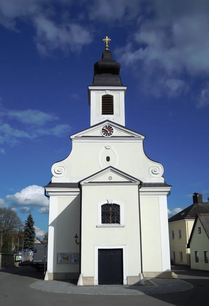 Eine weiße Kirche mit einem Turm und einer Uhr steht unter einem blauen Himmel. Sie hat ein schwarzes Dach, ein großes Rundbogenfenster und eine schwarze Tür.