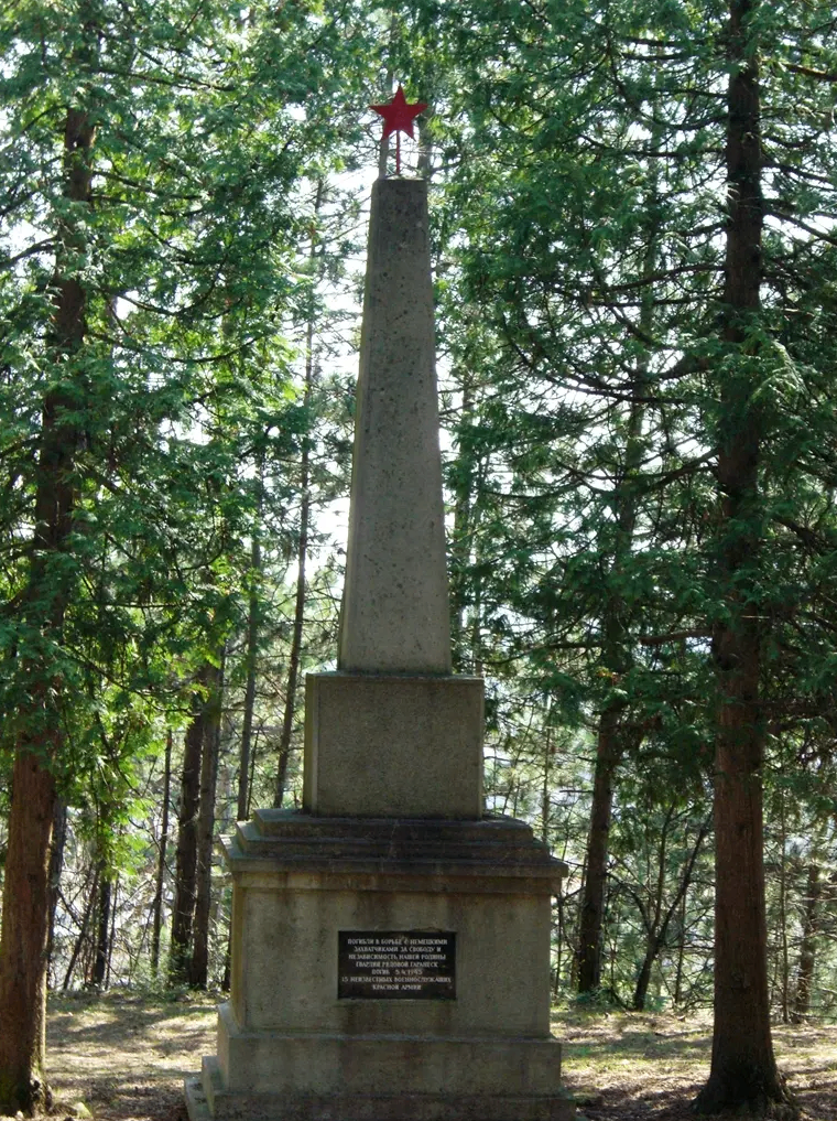 Ein hoher Steinobelisk mit einer Inschrift steht in einem Wald, umgeben von hohen Bäumen. Der Wald ist dicht, mit Sonnenlicht, das durch die Blätter fällt.