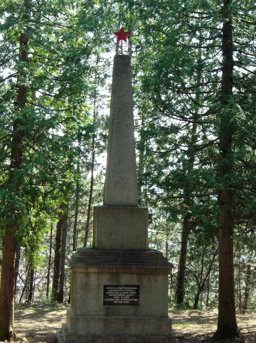 Ein hoher Steinobelisk mit einer Inschrift steht in einem Wald, umgeben von hohen Bäumen. Der Wald ist dicht, mit Sonnenlicht, das durch die Blätter fällt.