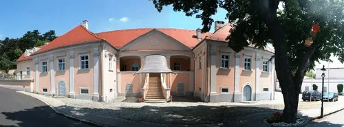 Ein großes rosa Gebäude mit rotem Dach und mehreren Fenstern, eine Treppe zur Eingangstür, ein Baum davor und geparkte Autos an der Seite.