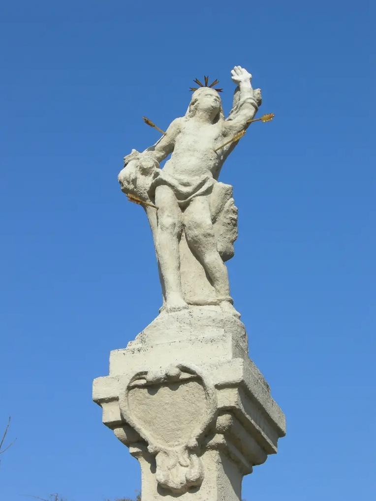 Eine weiße Statue eines Mannes mit goldener Pfeilkrone, der einen Bogen und einen Pfeil hält, steht auf einem Steinsockel unter einem klaren blauen Himmel.