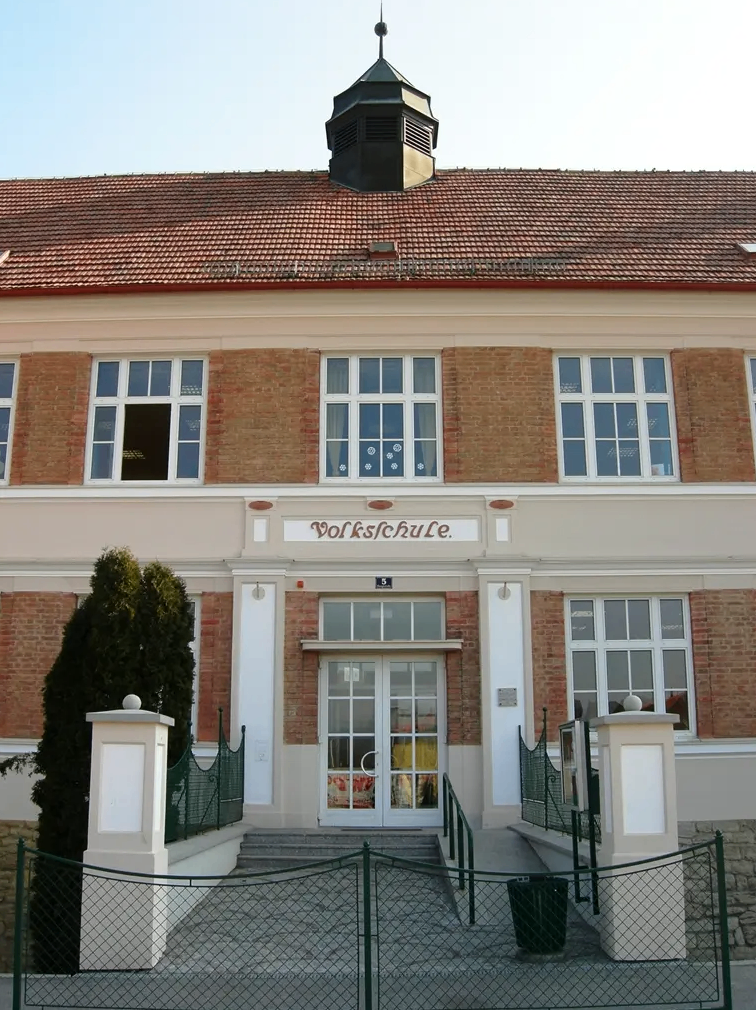 Die Vorderseite eines Ziegelgebäudes mit Glasfenstern und Türen, einem Schornstein oben und einem Schild, das 'Volksschule' lautet.
