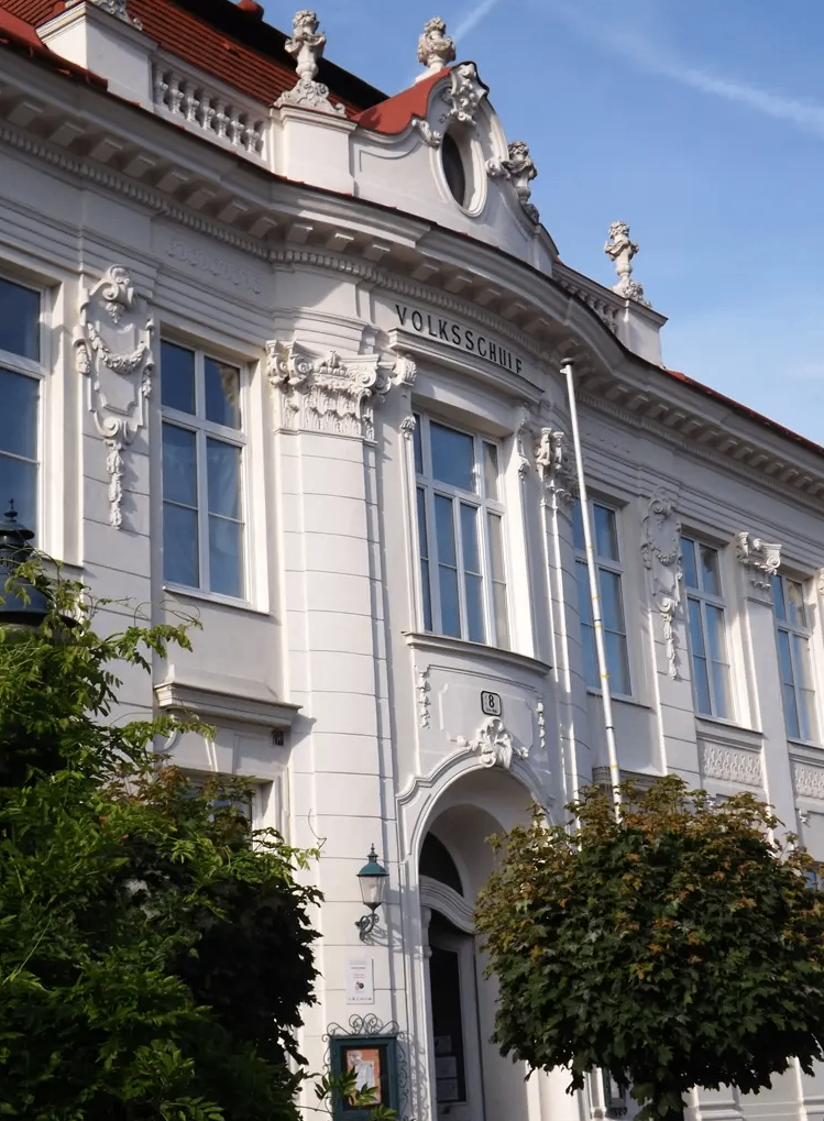 Ein weißes Gebäude mit verzierten Details und Glasfenstern. Es hat einen Haupteingang mit einem Bogen und einer Lampe an der Seite. Der Name 'Volksschule' steht über dem Eingang.