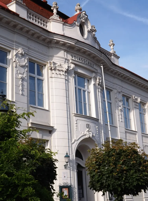 Ein weißes Gebäude mit verzierten Details und Glasfenstern. Es hat einen Haupteingang mit einem Bogen und einer Lampe an der Seite. Der Name 'Volksschule' steht über dem Eingang.