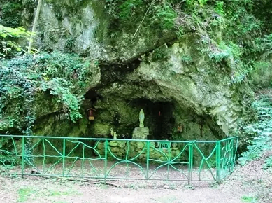 Eine kleine Grotte mit einem grünen Metallzaun, mit einer Statue eines Heiligen und einem Steinaltar im Inneren, umgeben von üppiger Grünfläche und Pflanzen.
