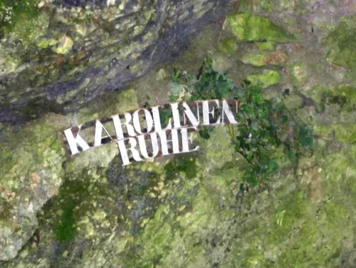 Eine Nahaufnahme einer moosbewachsenen Felswand mit den weiß geschriebenen Worten 'KAROLINEN RUHE'. Der Felsen hat eine raue Textur.