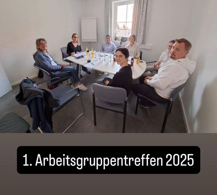 Eine Gruppe von Menschen sitzt in einem Besprechungsraum um einen Tisch. Eine Frau schreibt auf Papier. Flaschen, Stifte und Papiere liegen auf dem Tisch. Eine weiße Tafel und ein Fenster mit Vorhängen sind an der Wand.