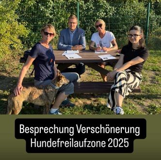 Vier Personen sitzen an einem Picknicktisch mit einem Hund in der Nähe. Papiere und Stifte sind auf dem Tisch, und sie diskutieren etwas. Das Bild hat die Worte 'Besprechung Verschönerung Hundefreilaufzone 2025'.