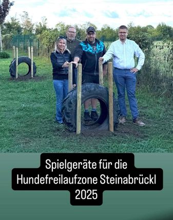 Vier Personen posieren neben einem Reifenhindernis in einer Grasfläche. Sie stehen um eine Holzstruktur herum. Ein Mann hat seine Hand auf der Hüfte.