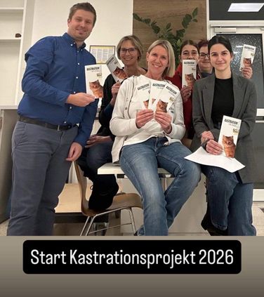Eine Gruppe lächelnder Menschen hält Bücher und Papiere mit Katzen darauf in den Händen. Sie stehen und sitzen in einem Raum. Ein Wasserzeichen lautet Start Kastrationsprojekt 2026.