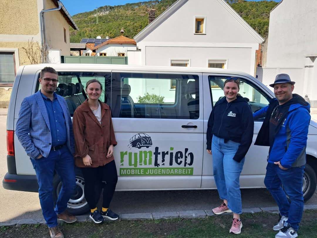 Vier Personen stehen neben einem weißen Van mit den Aufschriften 'rumtrieb' und 'MOBILE JUGENDARBEIT'. Sie lächeln und scheinen für ein Foto zu posieren. Dahinter befinden sich zwei Häuser und ein Hügel im Hintergrund.