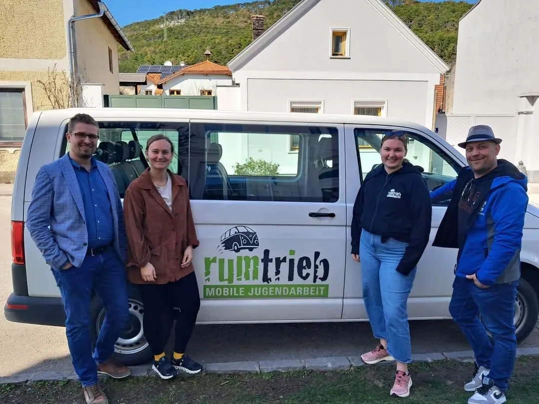 Vier Personen stehen neben einem weißen Van mit den Aufschriften 'rumtrieb' und 'MOBILE JUGENDARBEIT'. Sie lächeln und scheinen für ein Foto zu posieren. Dahinter befinden sich zwei Häuser und ein Hügel im Hintergrund.