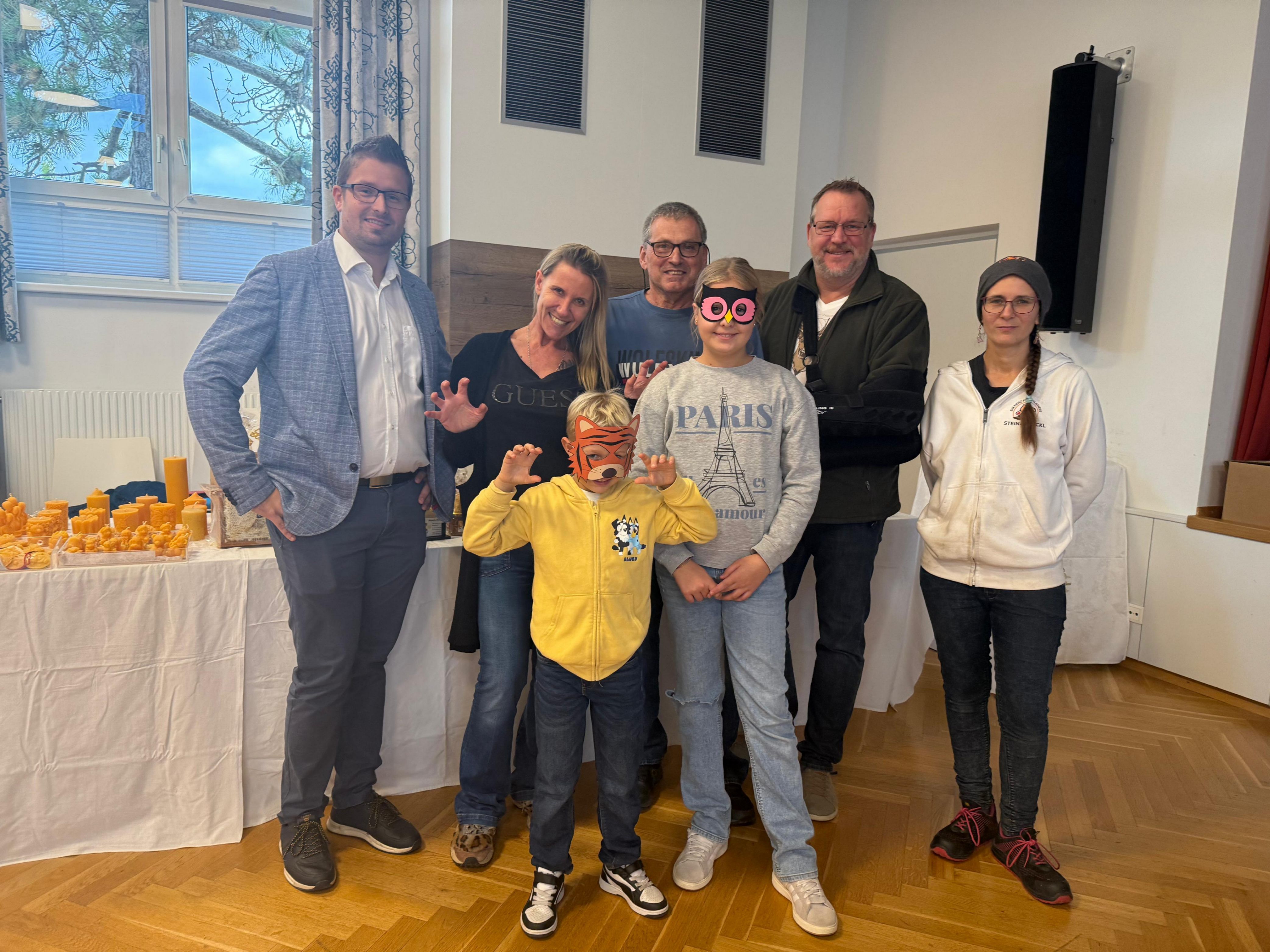Eine Gruppe von Erwachsenen und Kindern steht drinnen. Einige tragen eine Brille, Masken und Turnschuhe. Hinter ihnen steht ein Tisch mit Gegenständen.