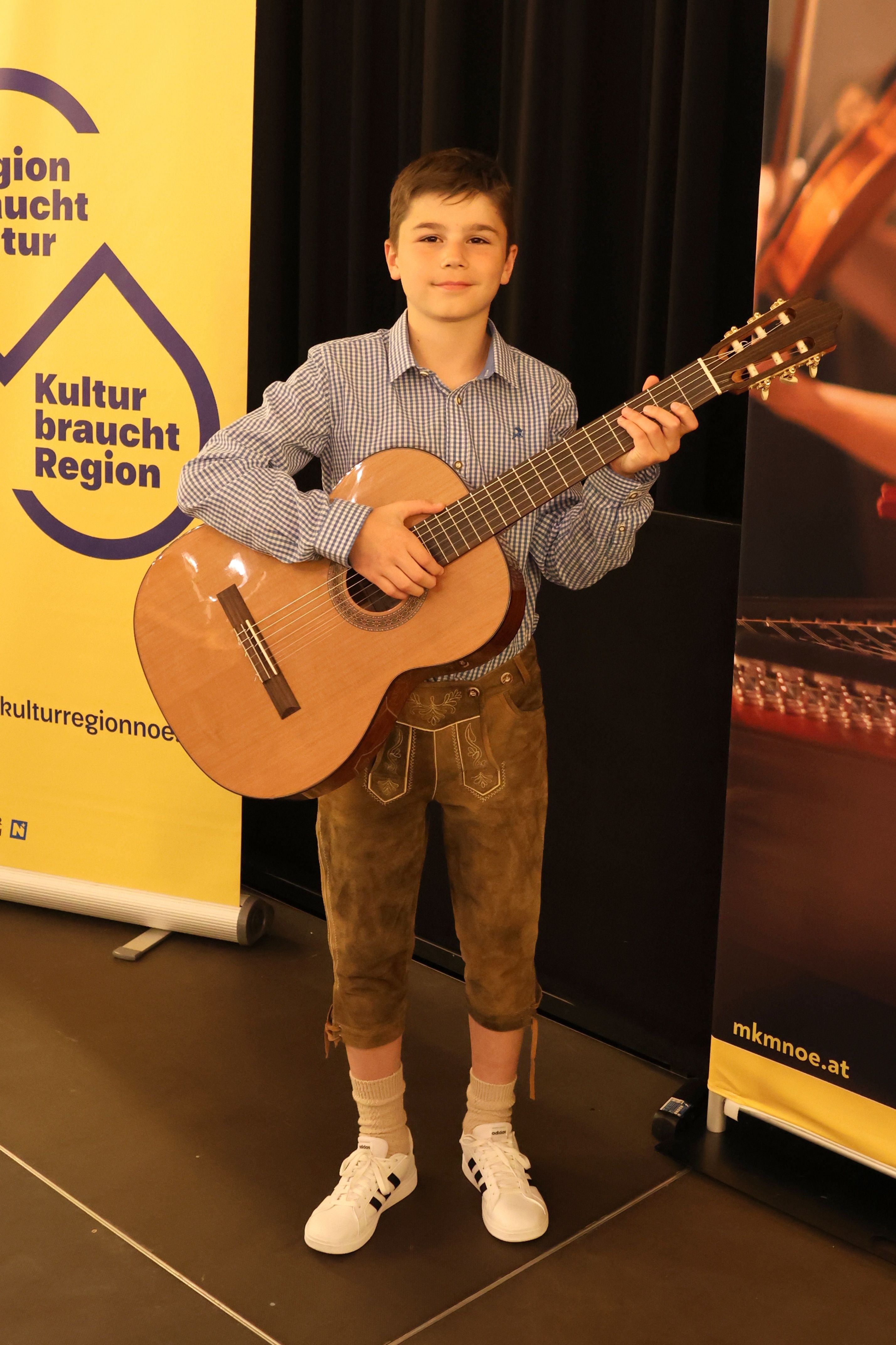 Ein Junge in traditioneller bayerischer Kleidung hält eine klassische Gitarre vor einem gelben Banner.