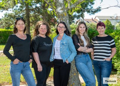 Fünf Frauen stehen lächelnd in einer Reihe, eine Frau lehnt sich an einen Baumstamm. Hinter ihnen stehen Bäume und ein Haus.