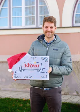 Ein Mann lächelt, während er eine Box mit der Aufschrift 'Advent in Hornstein' hält, trägt einen Weihnachtsmannhut und eine grüne Jacke. Er steht draußen vor einem Gebäude mit Fenstern.