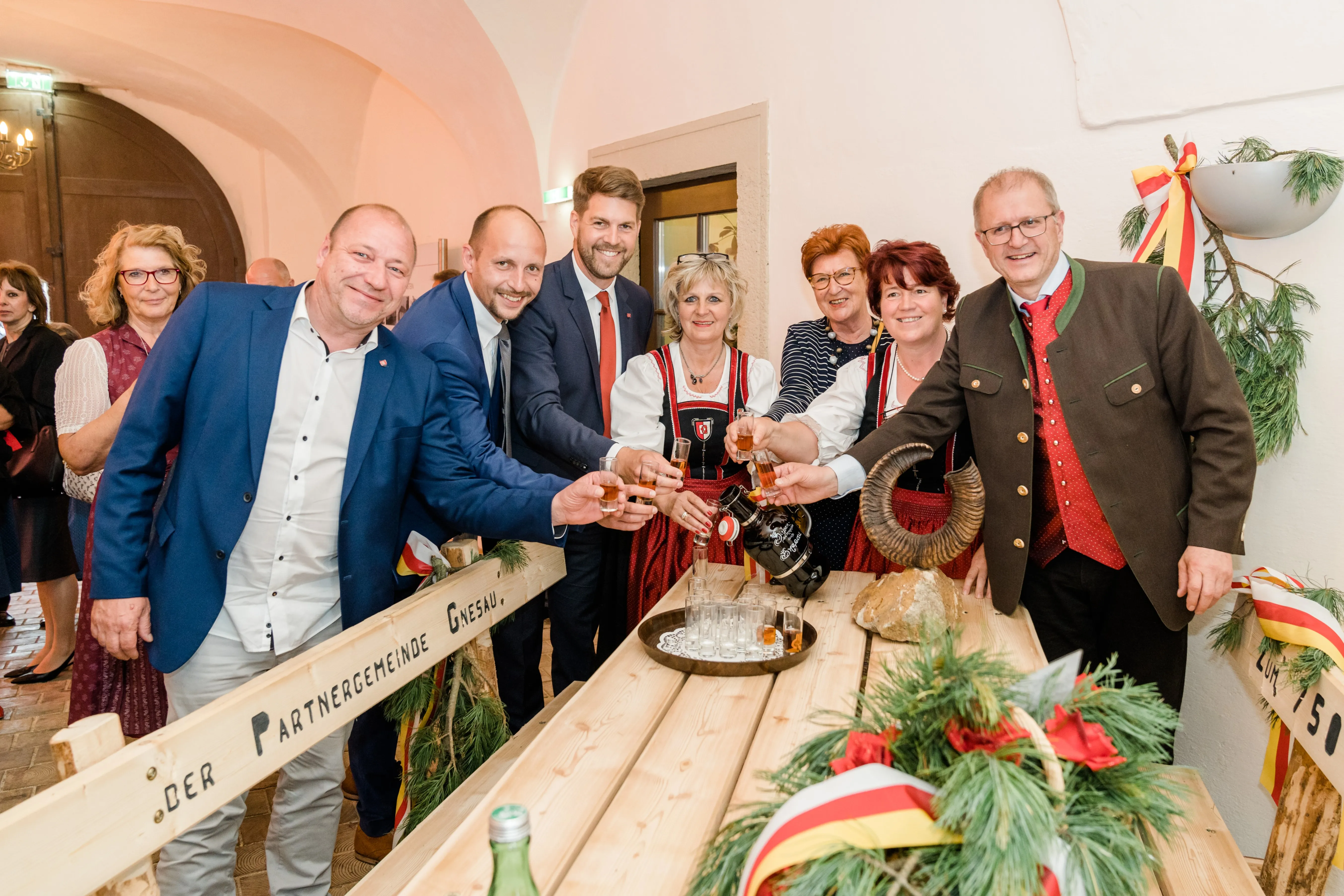 Eine Gruppe von Menschen lächelt und hebt Gläser mit Getränken auf einem Holztisch. Sie sind in formeller Kleidung gekleidet und einige tragen traditionelle deutsche Kleidung. Ein großer Horn ist auf dem Tisch und es gibt festliche Dekorationen.