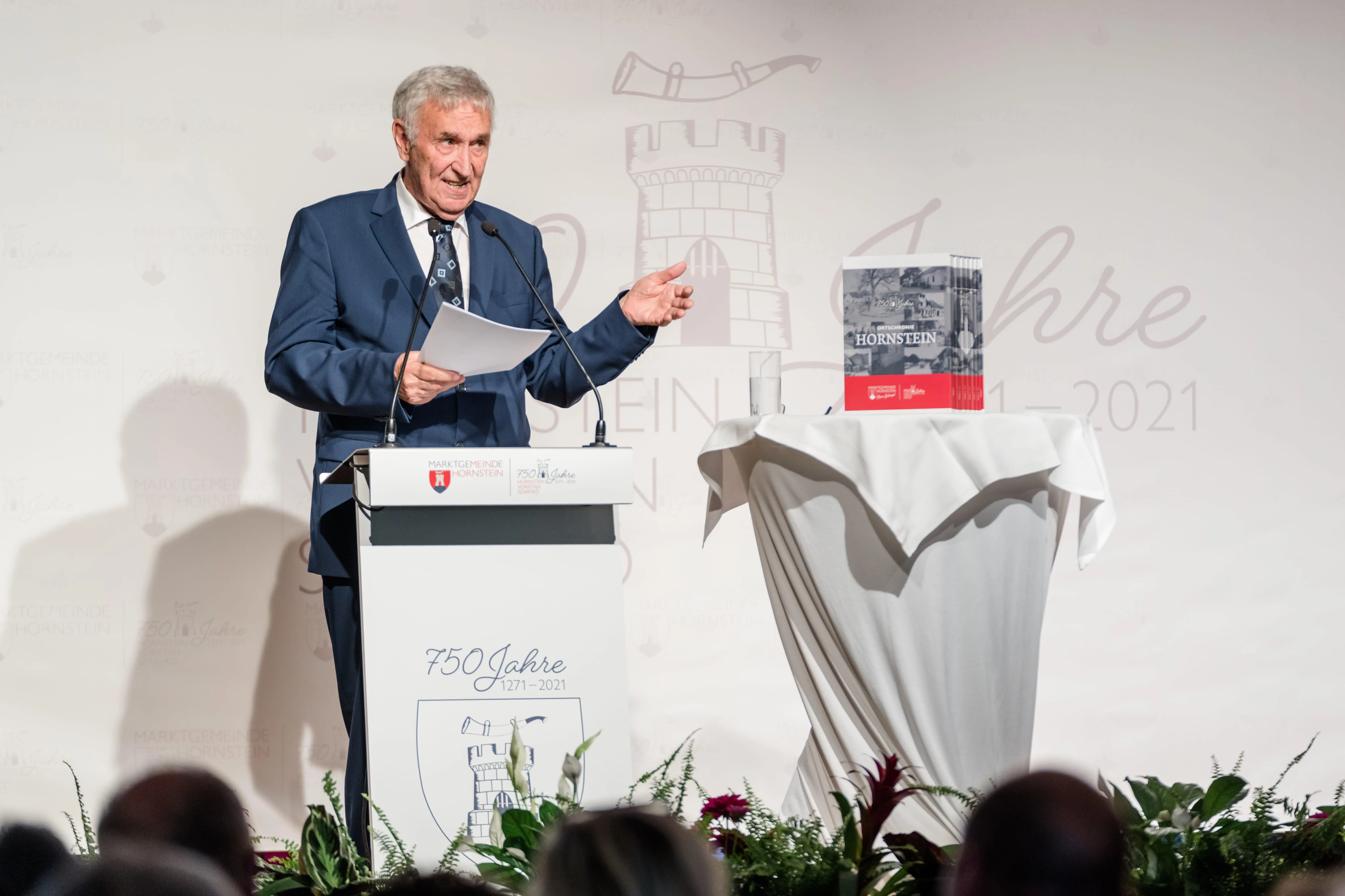 Ein Mann steht an einem Rednerpult, hält ein Papier in der Hand, lächelt, mit einem Mikrofon und Büchern auf dem Tisch dahinter, vor einer dekorierten Wand und einem Publikum.