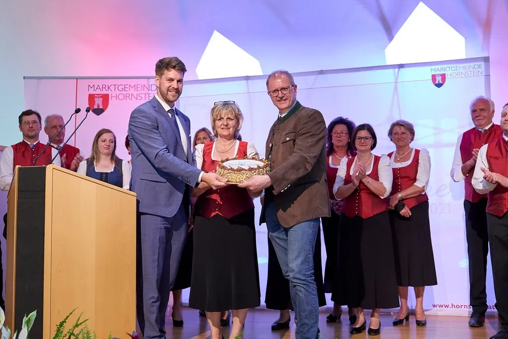 Ein Mann und eine Frau überreichen einem anderen Mann ein goldfarbenes Objekt bei einer Veranstaltung. Sie stehen an einem Podium, während mehrere Frauen in traditioneller Kleidung applaudieren. Die Umgebung ist mit einer violetten Wand mit einem Logo ausgestattet.