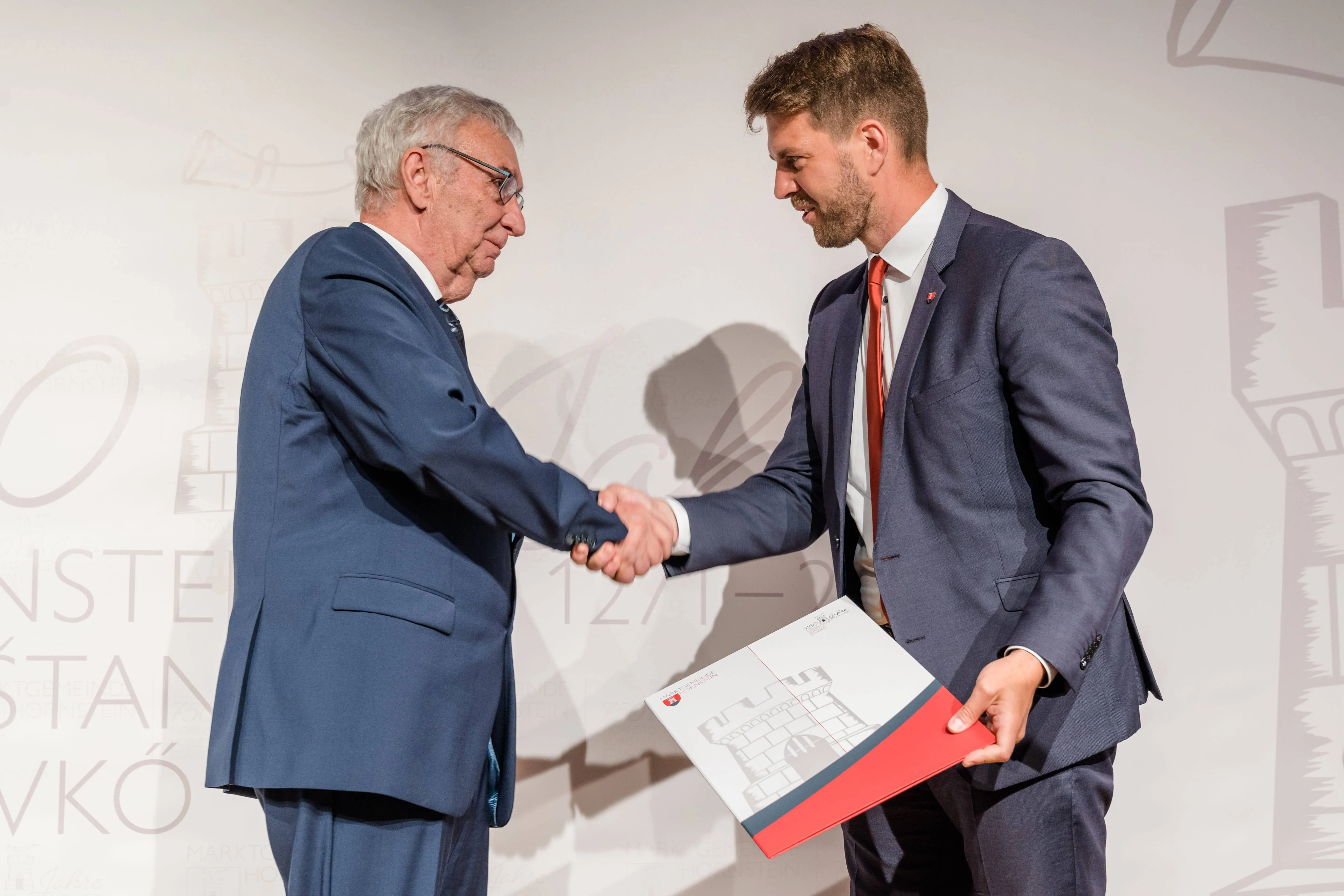 Ein älterer Mann mit grauem Haar drückt einem jüngeren Mann in einem blauen Anzug die Hand. Der ältere Mann hält ein Zertifikat. Der jüngere Mann hält ein Dokument.
