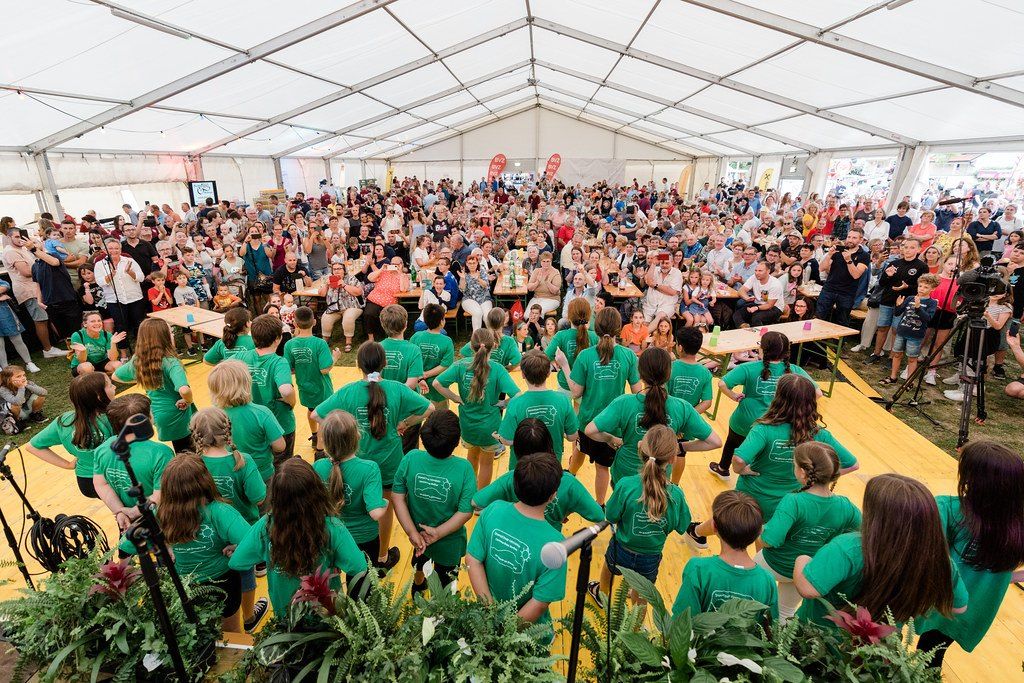 Eine große Menschenmenge in einem Zelt beobachtet eine Gruppe von Kindern in grünen T-Shirts, die auf der Bühne singen.