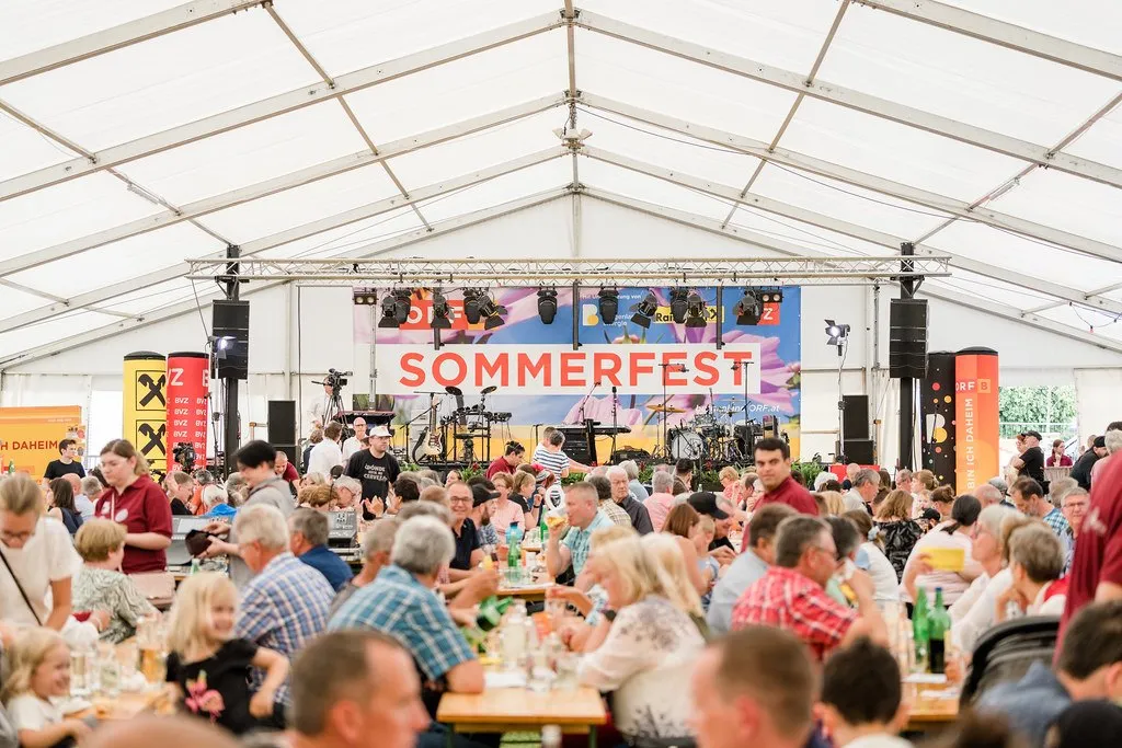 Eine Menschenmenge sitzt in einem großen Zelt an Tischen. Im Hintergrund befindet sich eine Bühne mit einem Banner, auf dem 'Sommerfest' steht. Musikinstrumente, Lautsprecher und Beleuchtungsgeräte sind aufgebaut.