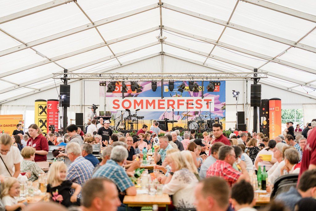 Eine Menschenmenge sitzt in einem großen Zelt an Tischen. Im Hintergrund befindet sich eine Bühne mit einem Banner, auf dem 'Sommerfest' steht. Musikinstrumente, Lautsprecher und Beleuchtungsgeräte sind aufgebaut.