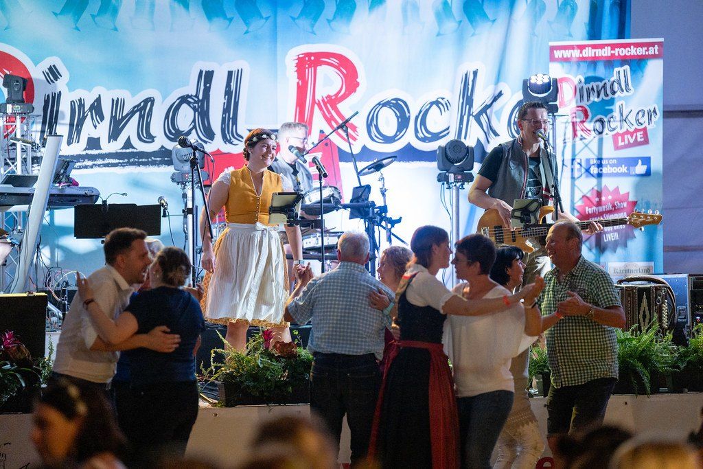 Eine Band spielt auf der Bühne mit einem großen Banner, das 'Dindl Rock' steht. Eine Frau in einem gelben Kleid singt, während ein Mann Gitarre spielt. Mehrere Menschen tanzen auf der Bühne.