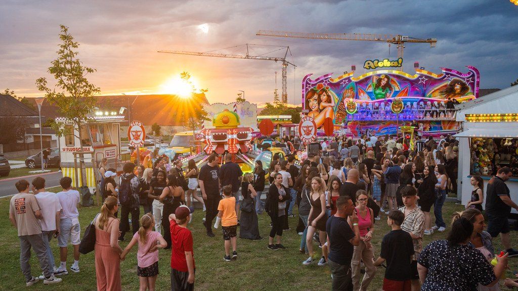 Eine Menschenmenge versammelt sich auf einem Jahrmarkt mit Fahrgeschäften, Ständen und einem Sonnenuntergang im Hintergrund. Die Leute unterhalten sich und genießen die Atmosphäre.