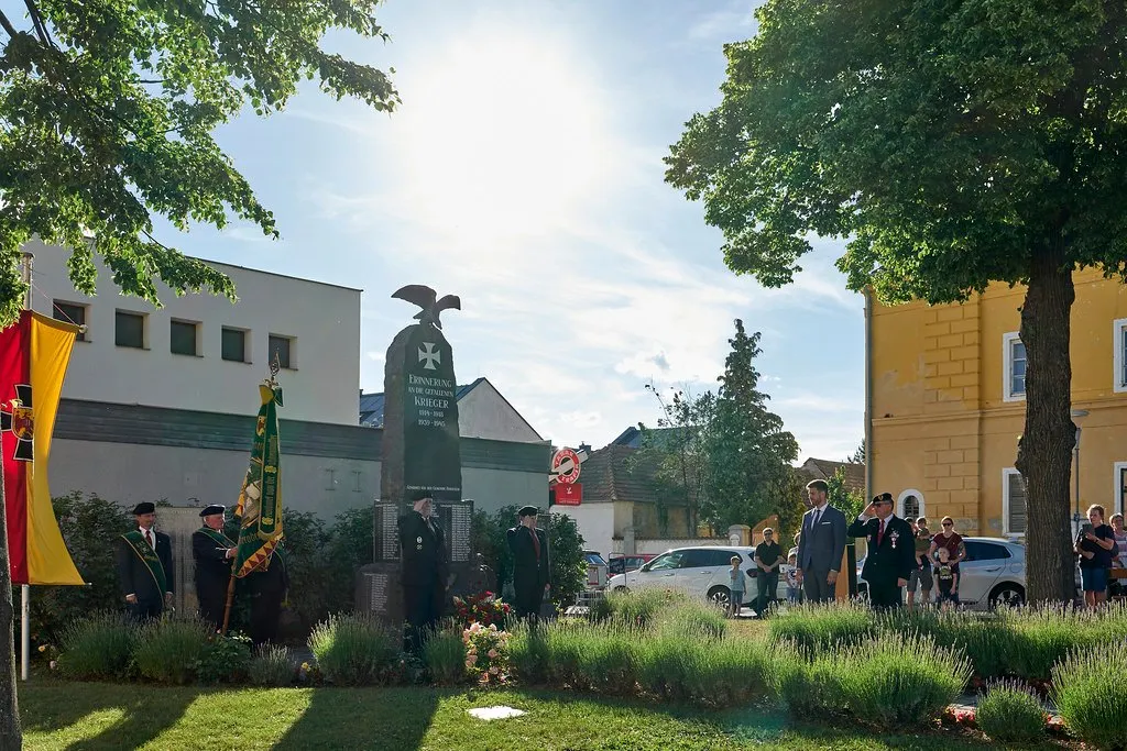 Ein Denkmal mit einer Adlerstatue und einer Flagge steht in einem Garten. Menschen in Uniformen stehen darum herum, einige salutieren. Ein Auto und ein Haus sind im Hintergrund.
